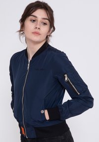Giacca bomber navy in tessuto liscio, con chiusura zip, tasche laterali e colletto e polsini a costine. Include una tasca con zip a contrasto.