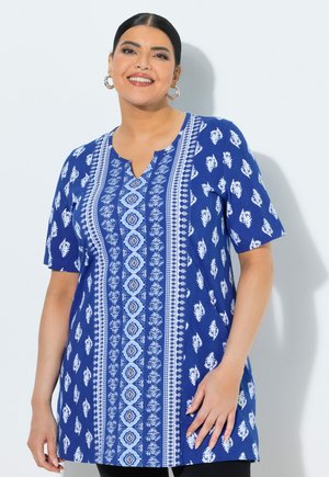 Lachende vrouw met donker haar, met grote oorbellen, draagt een blauw kort mouwig tuniek met witte bloem- en geometrische patronen.