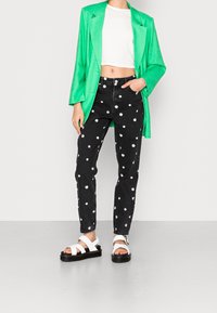 Blazer de satén verde sobre un top corto blanco, combinado con jeans negros con patrones de margaritas blancas y sandalias gruesas blancas.
