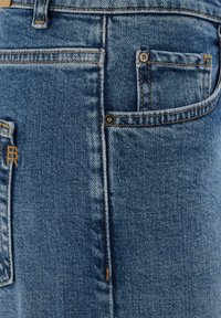 Približnji posnetek modrih kavbojk iz jeansa, ki prikazuje sprednji žep z majhnim žepkom za kovance, kovinske zakovice in natančno šivanje.