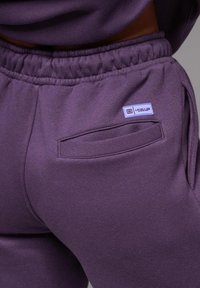 Fialový sweatshirťový materiál s elastickým pasem, boční kapsou a malou nášivkou "Level Up" na zadní straně. Měkká textura.