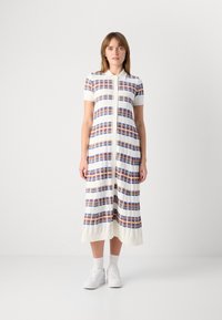  Γυναίκα που στέκεται φορώντας ένα λευκό midi φόρεμα με πορτοκαλί και μπλε οριζόντιες ρίγες, κοντά μανίκια, λευκά αθλητικά παπούτσια και κάλτσες.