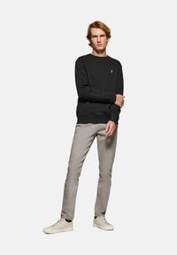 Svart sweatshirt med rund halsringning, ribbad ärm- och nederkant. Kombinerad med ljusgrå byxor och beige sneakers, stående mot en enfärgad bakgrund.