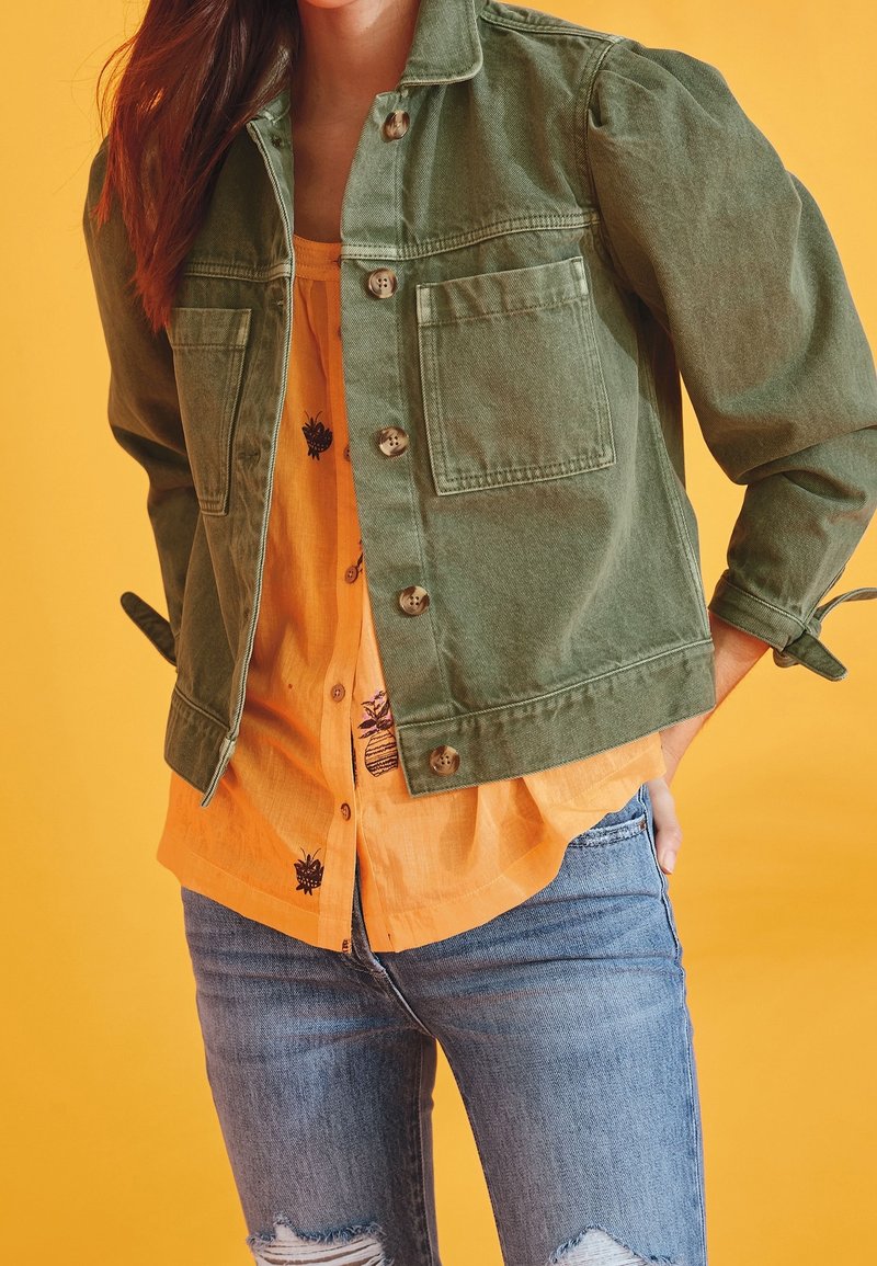 Femme portant une veste boutonnée vert olive sur une chemise orange imprimée et un jean bleu déchiré sur un fond jaune.