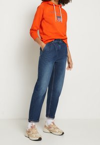 Pomarańczowa bluza z kapturem z logo, niebieskie dżinsy o luźnym kroju oraz beżowe trampki z różowymi akcentami. Materiały z bawełnianej mieszanki, styl casual.