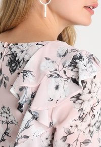 Blusa arricciata con motivo floreale in bianco e nero su sfondo rosa; presenta una chiusura con zip sul retro e orecchini ad anello argentati.