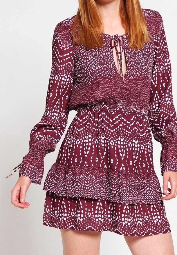 Bordeaux farvet kjole med lange ærmer, elastisk talje og lagdelt ruffle-kant. Har et hvidt abstrakt mønster og en bindemulighed ved halsudskæringen.