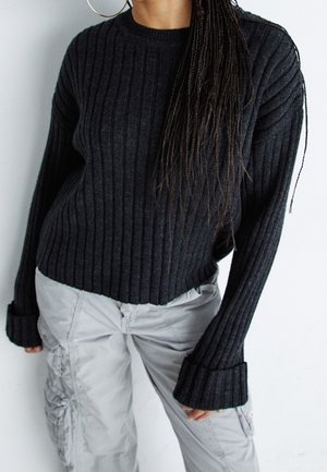 Person iført en mørk ribbet sweater med lange manchetærmer og lysegrå cargobukser, stående mod en ensfarvet hvid baggrund.