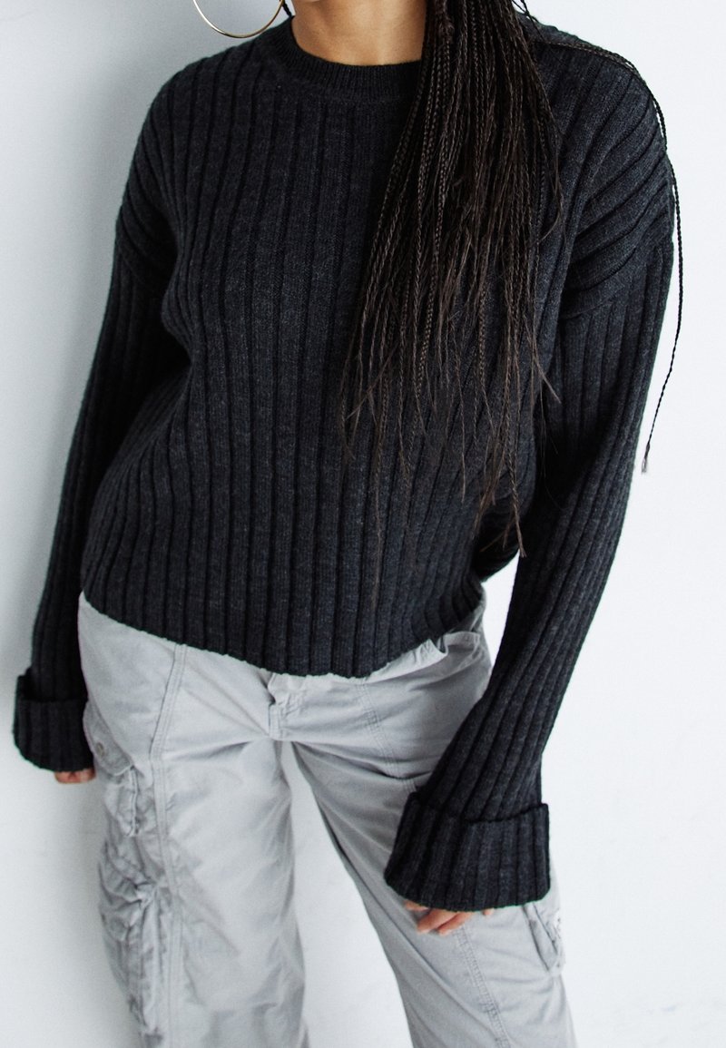 Personne portant un pull côtelé sombre avec des manches longues retroussées et un pantalon cargo gris clair, debout devant un fond blanc uni.