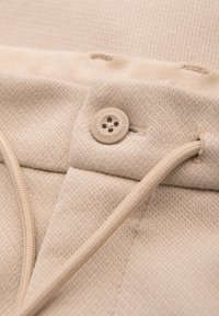 Ceinture en tissu texturé beige avec un bouton marqué « OMBRE » et un cordon assorti passé dans un œillet.