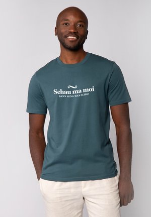 Teal T-shirt met witte tekst "Schau ma moi DANN SENG MAS SCHÓ!" Ronde hals en korte mouwen, gemaakt van gladde stof, gedragen met lichte broek.