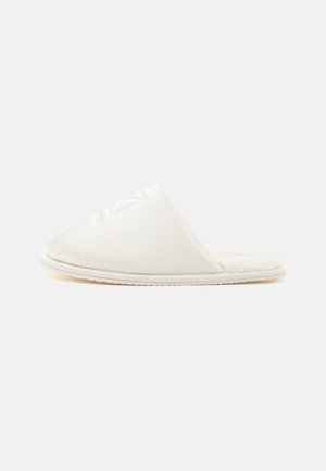 Zapatilla blanca tipo slip-on con parte superior acolchada y forro interior suave y peludo, mostrada de lado sobre un fondo liso.