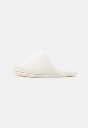 Zapatilla blanca tipo slip-on con parte superior acolchada y forro interior suave y peludo, mostrada de lado sobre un fondo liso.