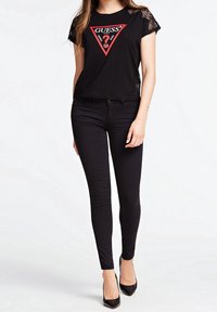 Mujer vestida con una camiseta Guess de manga de encaje negra y pantalones ajustados negros, con tacones de punta negra, de pie frente a un fondo blanco.