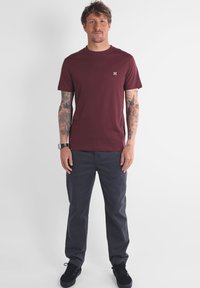 T-shirt en coton bourgogne avec col rond, porté avec un pantalon gris foncé. Présente un petit logo sur la poitrine et des tatouages visibles sur les bras.