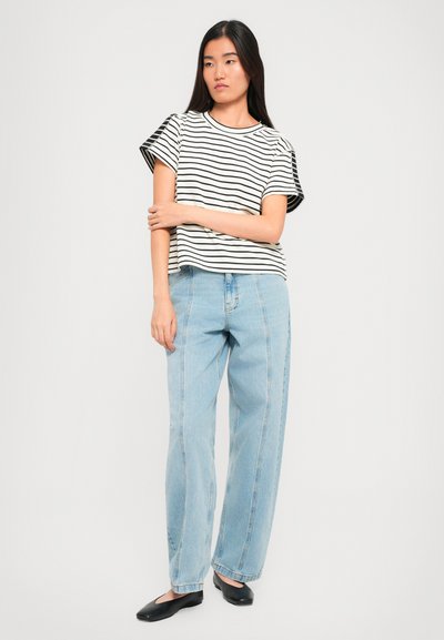 Young woman with long black hair wears a short-sleeve black-and-white striped shirt, light blue jeans, and black flats, standing against white background.