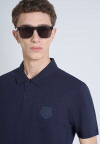 Mornarica polo majica s teksturiranim logom medvjeda na lijevoj strani grudi, s klasičnom kragnom i tri gumba, u kombinaciji s tamnim sunčanim naočalama.