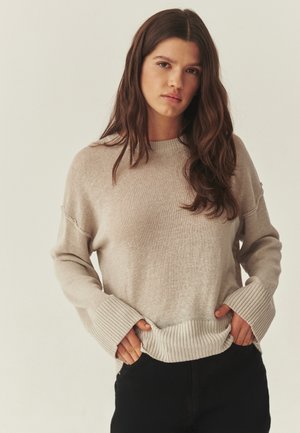 Mladá žena s dlhými hnedými vlasmi, ktorá má na sebe béžový pletený sveter a čierne nohavice, ruky zasunuté do lemu svetra, neutrálny pozadie.