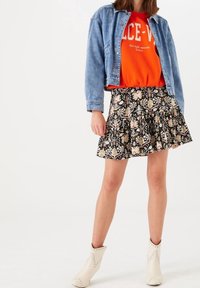 Džínová bunda, oranžová mikina s potiskem, černá květovaná plisovaná sukně, kotníkové boty s cvočky, celkový ležérní vrstvený outfit.