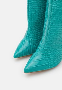 Botas de tacón alto turquesa hechas de cuero texturizado con estampado de cocodrilo, que presentan una punta afilada y un acabado suave con detalles de costura visibles.