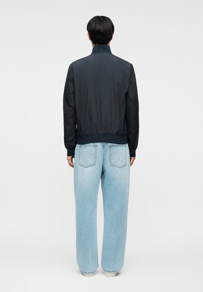 Άτομο που στέκεται πλάτη, φορώντας μαύρο μπουφάν και ανοιχτόχρωμα μπλε φαρδιά τζιν, σε απλό φόντο.