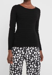 Schwarzes Langarmoberteil mit strukturiertem geripptem Design, rundem Ausschnitt und taillierter Form, kombiniert mit schwarz-weißen gemusterten Hosen.