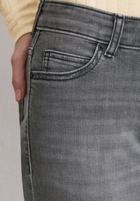 Gros plan sur la main d'une personne reposant à côté de la poche d'un jean en denim gris, portant un haut en maille côtelée beige clair à manches longues.