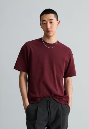 Jeune homme aux cheveux courts portant un t-shirt bordeaux et un pantalon noir à cordon, debout les mains dans les poches devant un fond uni.
