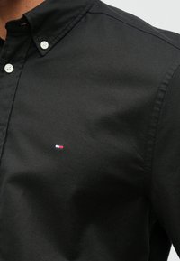 Čierna košeľa na gombíky s špicatým golierom, vyrobená z hladkej látky. Má malé vyšívané logo v červenej, bielej a modrej farbe na hrudi.