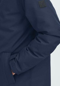 Mornarsko modra jakna z gladko, mat površino. Ima logotip na patchu na rokavu in stranski žep. Preprost, funkcionalen dizajn.