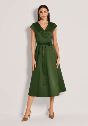 Frau steht und trägt ein grünes Midi-Kleid mit V-Ausschnitt, kurzen Ärmeln, gebundenem Taillengürtel und braunen spitzen Schuhen vor einem schlichten Hintergrund.