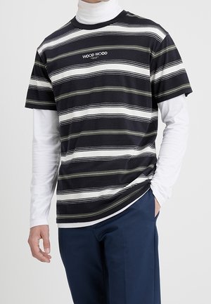Mężczyzna w czarno-białej pasiastym t-shircie Wood Wood na białej koszulce z długim rękawem, w granatowych spodniach, z ręką w kieszeni.
