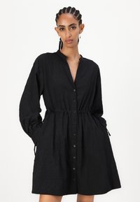 Rochie neagră cu mâneci lungi, închidere cu nasturi și talie elastică. Material texturat, cu o croială lejeră și buzunare laterale.