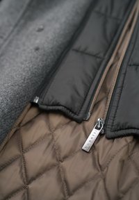 Gepolsterte schwarze Jacke mit Reißverschluss, ausgestattet mit einem gesteppten braunen Futter und einem silbernen Metallreißverschluss mit dem Markennamen.