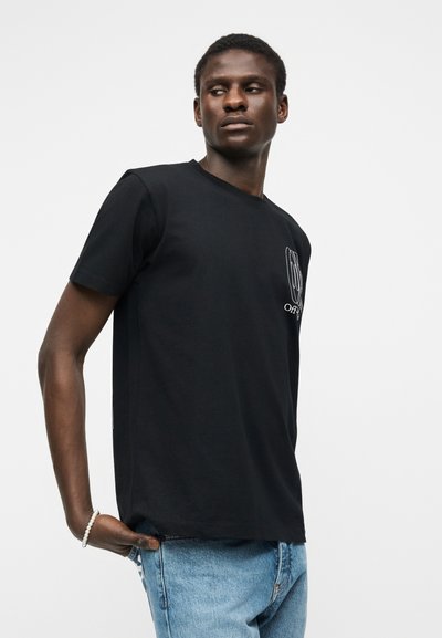 Άνδρας που φοράει μαύρο μπλουζάκι με σχέδιο και ανοιχτόχρωμο τζιν, κοιτάζει πλάγια με το ένα χέρι στην τσέπη, σε λευκό φόντο.