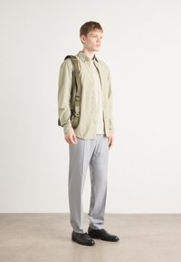 Conjunto masculino que incluye una camisa de botones verde claro, una parte superior crema, pantalones grises y zapatos negros. Incluye un bolso de hombro con correa ajustable.