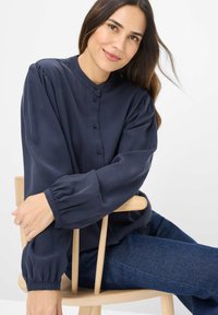 Mørkeblå, langærmet bluse med høj hals, knaplukning og samlede ærmer, lavet af et glat stof. Model siddende på en træstol.