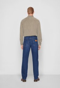 Hombre de pie de espaldas, vestido con una camisa tostada, jeans oscuros con bolsillos traseros y zapatos de cuero marrón, sobre un fondo liso.