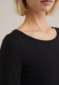 Fekete bordás, hosszú ujjú felső, széles kerek nyakkivágással. Az anyag puhanak és testhezállónak tűnik, hangsúlyozva a váll és a kulcscsont környékét.