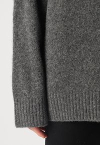 Pull en laine gris présentant une texture douce, des poignets côtelés et une coupe décontractée, photographié du coude vers le bas sur un fond uni.