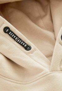 Felpa beige con cappuccio, caratterizzata da un'etichetta nera con testo bianco, realizzata in un tessuto morbido con una consistenza liscia e un design a V.