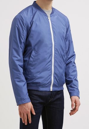 Mann trägt eine blaue Bomberjacke mit Reißverschluss über einem grauen T-Shirt und dunklen Jeans, steht vor einem schlichten hellen Hintergrund.