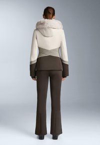 Eng anliegende Skijacke in Creme und Grau mit Kunstpelzkragen, kombiniert mit braunen ausgestellten Skihosen mit Reißverschluss an den Säumen.