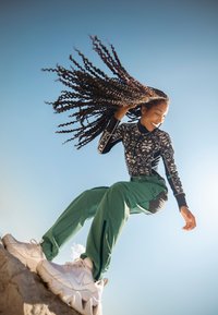 Tenue de sport comprenant un haut à manches longues noir et blanc à motifs, un pantalon vert et des baskets blanches imposantes. Ciel bleu vif en arrière-plan.
