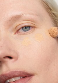 Fond de teint appliqué sur la peau, texture douce, couleur beige clair, petite éponge de maquillage en action, gros plan du visage avec des détails visibles de la peau.