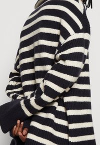 Pull rayé avec des couleurs alternées bleu marine et crème, texture côtelée, coupe ample avec manches larges et fentes latérales.