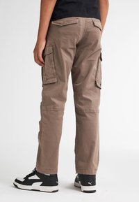 Cargo broek van lichtbruin stof met twee zijvakken, rechte pasvorm en versterkte stiksels. Draagt met zwart-witte sneakers.