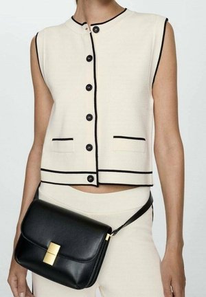 Kvinde iført ærmeløs cremefarvet top med sort kant og knapper, cremefarvede bukser og en sort læder crossbody-taske med guldlås.