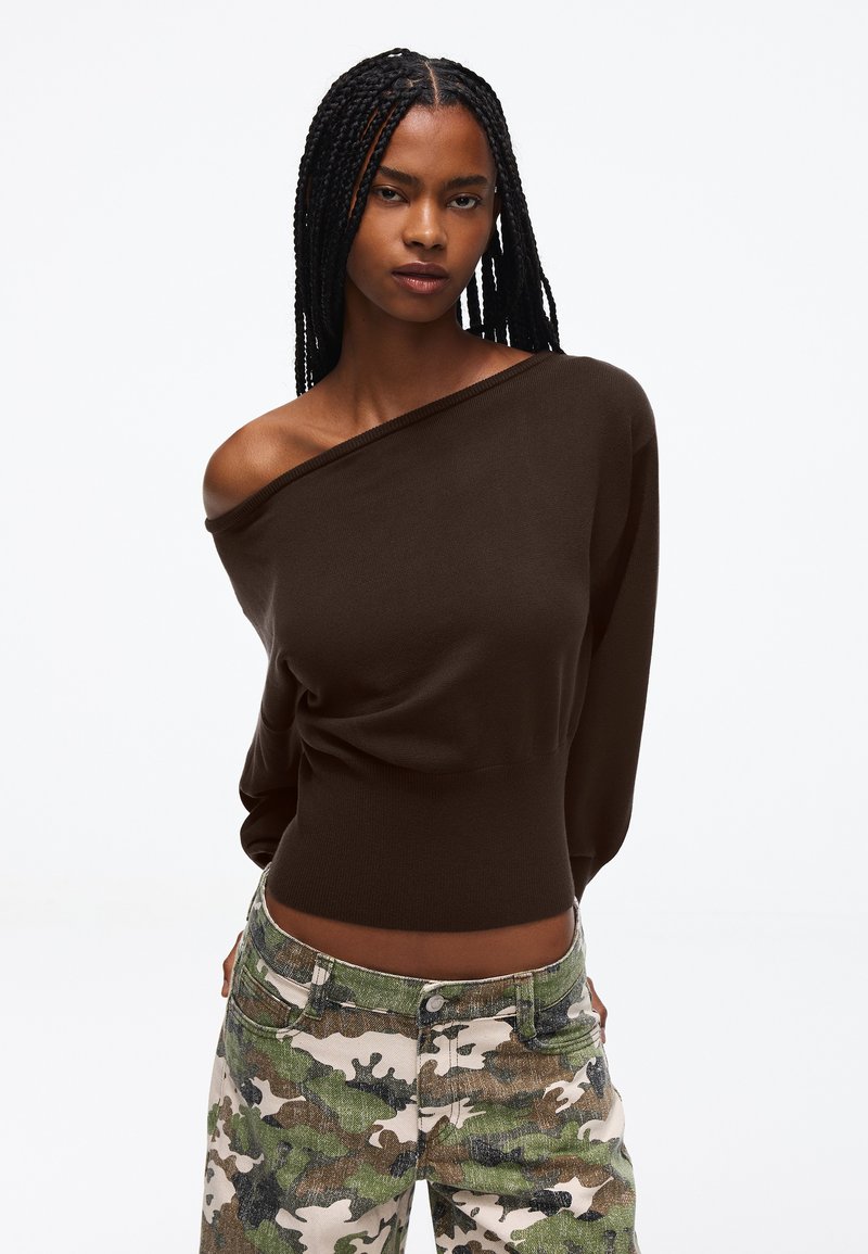 Jeune femme aux longues tresses portant un pull sombre épaules dénudées et un pantalon camouflage vert, debout devant un fond blanc uni.
