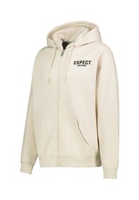 Beige zip-up hoodie with a front pocket, drawstring hood, and the text "EXPECT NOTHING" in black on the left side. Soft fabric texture.